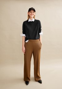 Schwarzer Strickpullover über einem weißen Hemd mit Kragen, kombiniert mit braunen weit geschnittenen Hosen und schwarzen spitzen Schuhen, vor einem neutralen Hintergrund.