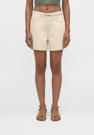 Donna che indossa pantaloncini beige con cintura, top corto in pizzo verde e sandali beige con cinturini, in piedi su uno sfondo chiaro e uniforme.