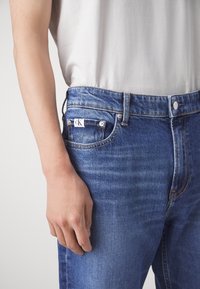 Light blue denim jeans with a straight cut, five pockets, and a CK logo tag on the back pocket. The fabric has a smooth texture.