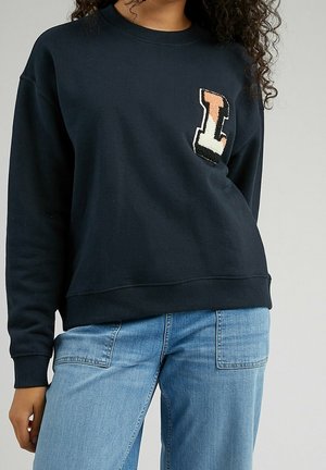 Frau trägt einen schwarzen Pullover mit einem strukturierten Patch-Buchstaben „L“ auf der Brust und hellblaue Jeans, steht vor einem einfarbigen Hintergrund.