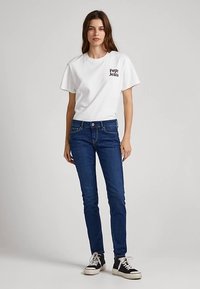 Fehér pamut póló fekete logóval, slim-fit kék farmerrel és fekete-fehér tornacipővel párosítva. Egyszerű kialakítás, hétköznapi fazon.