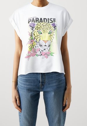 Chemise blanche sans manches avec manches retroussées présentant un visage de léopard coloré, des fleurs roses et le texte "Feeling Tropical PARADISE".