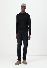 Čierny sveter s výstrihom do V, tmavé džínsy, béžové topánky; model má na hlave svetlošedú čiapku s textom. Jednoduchý, neformálny outfit na čistom pozadí.
