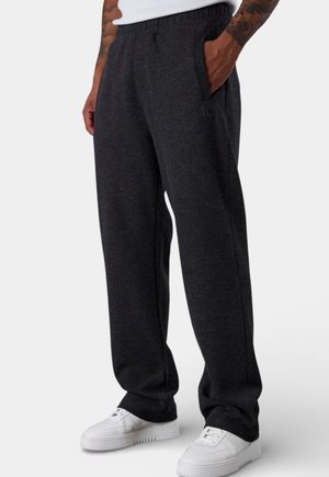 Mann trägt dunkelgraue Sweatpants in lockerer Passform und weiße Sneaker, steht mit einer Hand in der Tasche vor einem schlichten Hintergrund.