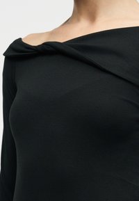 Κοντινό πλάνο ενός ατόμου που φοράει μαύρο τοπ μακρυμάνικο με ανοιχτούς ώμους και διπλωμένη λεπτομέρεια στον λαιμό.