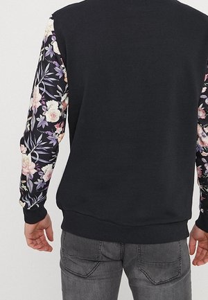 Mann trägt schwarzen Sweatshirt mit floral gemusterten Ärmeln und grauen Jeans, von hinten gesehen.