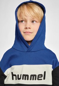 Niebieska bluza z kapturem z białą i czarną paskiem na klatce piersiowej, z nazwą marki "hummel" napisaną czarnym literami, wykonana z materiału bawełnianego.