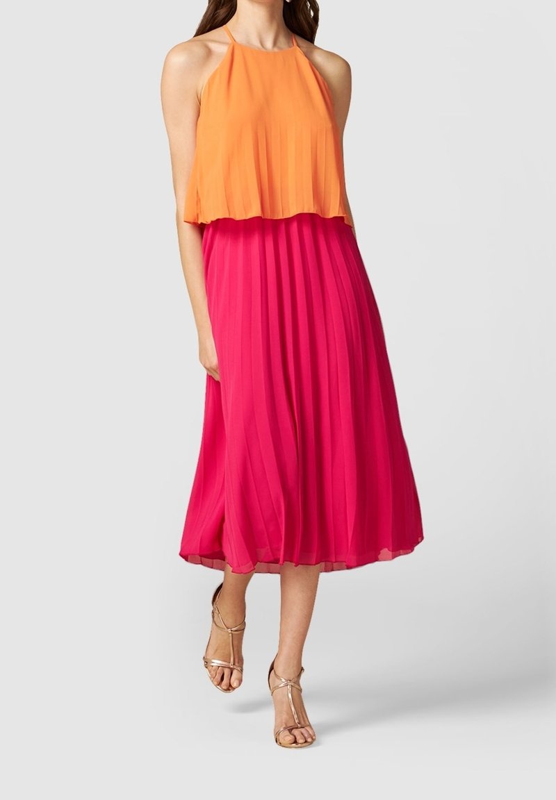 Femme portant un haut orange sans manches à col licou et une jupe midi plissée rose vif, avec des sandales à talons hauts beiges à brides.