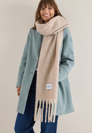 Sciarpa tessuta beige con una trama leggera e bordi frangiati, dotata di un piccolo etichetta. Indossata sopra un cappotto teal con un design sartoriale.