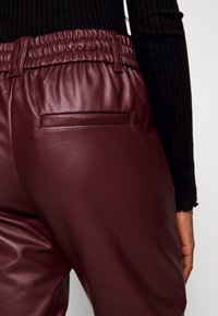 Gros plan sur une personne portant un pantalon en cuir bordeaux avec une ceinture élastique et une poche passepoilée, associé à un haut noir à manches longues côtelé.