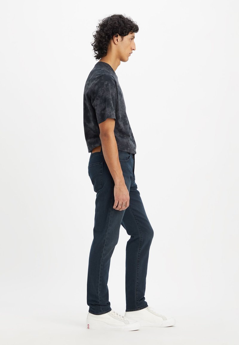 Chemise à manches courtes noir tie-dye, jean slim-fit bleu foncé, baskets blanches. Le mannequin se tient de profil devant un fond blanc uni.