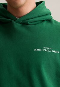 Man in een groene hoodie met witte tekst "HOSTED BY MARC O’POLO DENIM" op de borst, gecropt onder de neus.