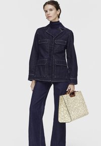 Chaqueta de denim con dos bolsillos frontales, lavado azul oscuro, combinada con pantalones de denim de pierna ancha. Sosteniendo un bolso crema con un patrón decorativo y asas marrones.