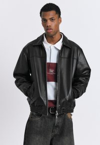 Mladý muž v černé kožené bundě přes bílou a bordó pruhovanou polokošili a tmavé džíny, stojící s rukama v kapsách.