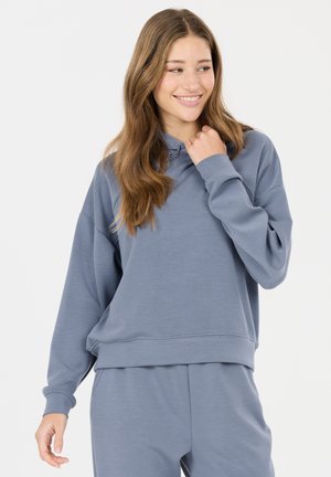 Jonge vrouw glimlachend, gekleed in een losse blauwe trui en bijpassende broek, kijkt naar haar rechterzijde tegen een effen witte achtergrond.