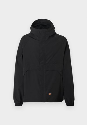 Juoda gobtuvu striukė su priekinio užtrauktuko uždarymu, elastingais rankogaliais, dviem užtrauktukais užsegamomis šoninėmis kišenėmis ir maža prekės ženklo logotipu prie apatinio krašto ant vienspalvio fono.