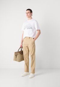 Hvit t-skjorte med grå grafikktrykk, beige chinos og hvite joggesko. Bringer en brun tote med teksturerte detaljer og et stilfullt design.