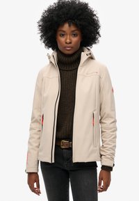 Beige softshell jack met een capuchon, rits aan de voorkant en oranje accenten. Draag het over een bruine gebreide coltrui en donkere jeans.