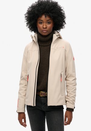 Beige softshell jacket with a hood, zippered front, and orange accents. Worn over a brown knitted turtleneck and dark jeans.