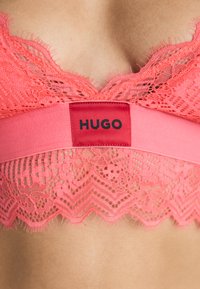 Korálový čipkový bralet s vlnitým okrajom a hladkým pásom s červenou nálepkou označenou "HUGO". Zložitá dekor�ácia a jemná textúra.