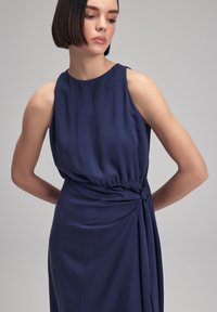 Navy blue sleeveless dress with a gathered waist and knot detail on the side. Smooth fabric with a slight sheen, featuring a round neckline.