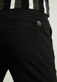 Zwarte broek met een gladde textuur, voorzien van een achterzak en een zichtbaar label. De stof lijkt lichtgewicht en op maat gemaakt.