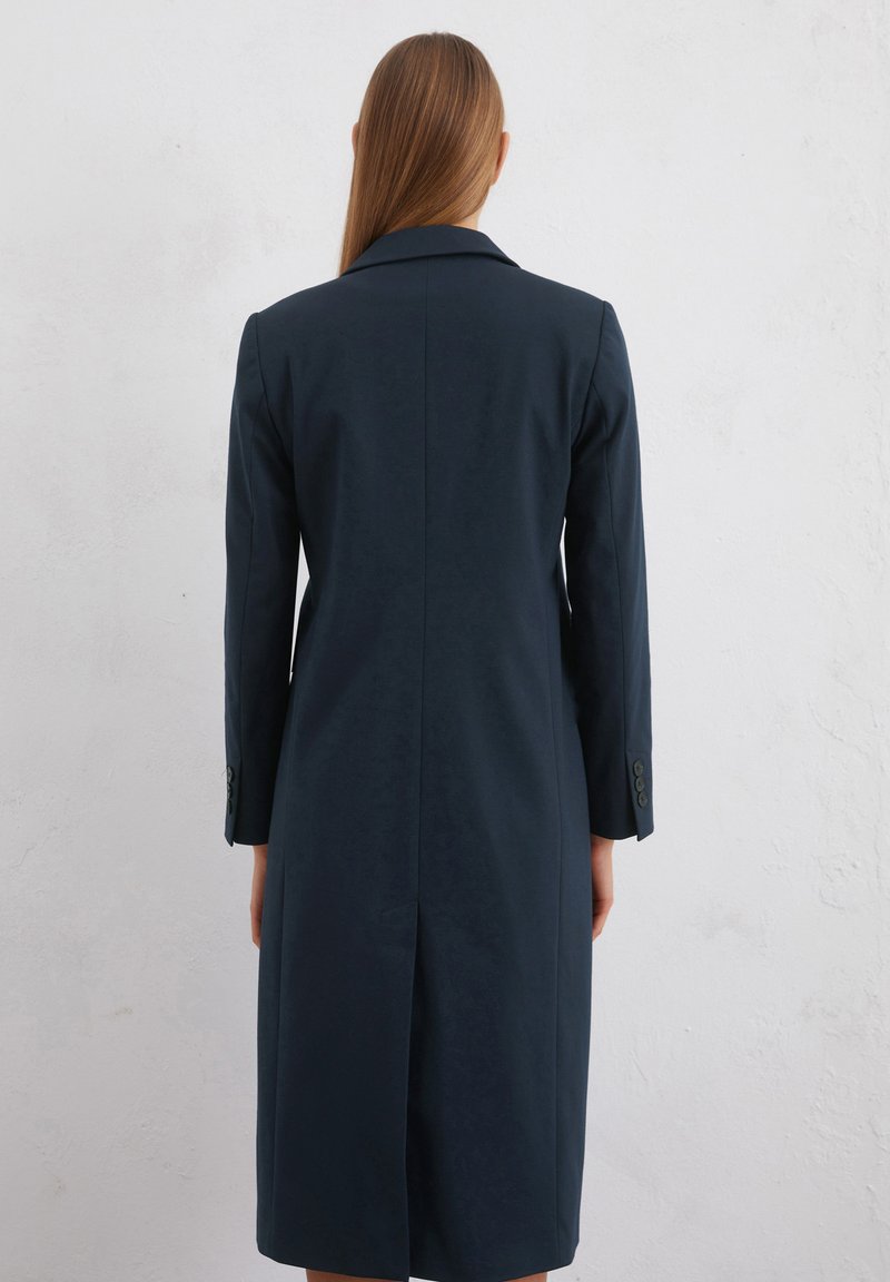 Marineblauer taillierter Mantel mit einem Schlitz hinten, spitzen Revers und geknöpften Manschetten an den langen Ärmeln. Glatte Stoffstruktur.