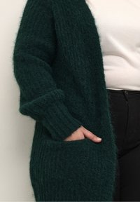 Grøn, fluffy strik cardigan med lange ærmer, ribbede manchetter og to sidelommer. Har en afslappet pasform og blød tekstur.