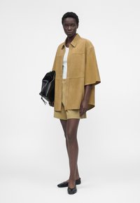 Γυναίκα που φοράει μια χακί oversized πουκάμισο με κουμπιά, ταιριαστό σορτς, λευκό τοπ, κρατά μια μαύρη τσάντα και μαύρα φλατ, στέκεται μπροστά σε απλό φόντο.