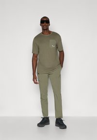 Ελαιόχρωμο κοντομάνικο μπλουζάκι με τσέπη στο στήθος και λογότυπο, συνδυασμένο με παντελόνι στο ίδιο χρώμα. Το μοντέλο φοράει μαύρα γυαλιά ηλίου και παπούτσια πεζοπορίας.