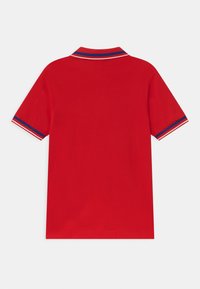 Červená polo košile s krátkým rukávem, s modrými, bílými a červenými pruhy na límci a manžetách rukávů, zobrazená ze zadní strany na bílém pozadí.