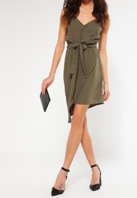 Oliivivihreä kietaisumekko, jossa on säädettävä solmuvyö, syvä V-pääntie ja polvenmittainen epäsymmetrinen helma. Yhdistetty mustien korkokenkien kanssa.