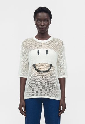 Νεαρή γυναίκα φορώντας λευκό τοπ από δίχτυ με μαύρο σχέδιο emoticon, συνδυασμένο με μπλε τζιν, στέκεται πάνω σε λευκό φόντο.