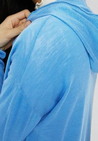 Jasnoniebieska bluza z kapturem o teksturowanej, prążkowanej powierzchni. Tkanina wydaje się miękka i nieco przezroczysta, z luźnym krojem i prostym wzorem.