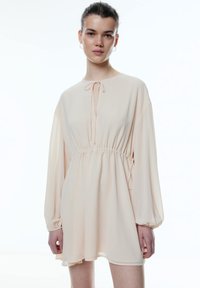 Kurzärmliges, blassrosa Kleid mit elastischem Bund und Bindeband am Hals. Verfügt über weit ausgestellte Ärmel und eine glatte, fließende Textur.