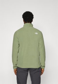 Veste en polaire verte avec un col haut, texture douce et logo blanc dans le haut du dos, dotée de manches longues et d'un ourlet droit.