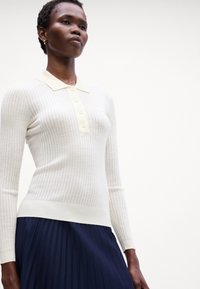 Fehér bordázott hosszú ujjú pólósweater gombos elülső résszel és világos sárga gallérral, amely navy kék redőzött szoknyával van párosítva.