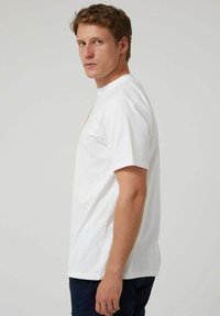 Camiseta de algodón blanca con mangas cortas y un escote redondeado, con un diseño gráfico en la parte frontal, mostrada en un modelo de perfil.