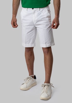 Uomo con pantaloncini bianchi fino al ginocchio, camicia verde e scarpe da ginnastica bianche, in piedi davanti a uno sfondo grigio uniforme.