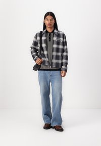 Black and white checkered flannel jacket over patterned black polo and light blue jeans. Black crossbody bag, brown shoes.