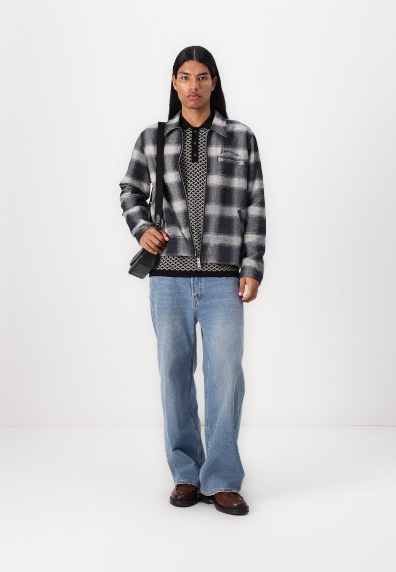 Schwarz-weißer karierter Flanelljacke über einem gemusterten schwarzen Poloshirt und hellblauen Jeans. Schwarze Umhängetasche, braune Schuhe.