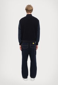Gilet in pile blu navy scuro con maniche blu, orlo a coste e etichetta gialla sui pantaloni. I pantaloni larghi presentano cuciture a contrasto e tasche.