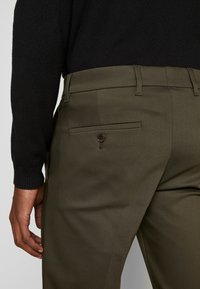 Pantaloni verde scuro realizzati in tessuto morbido, con una tasca posteriore con bottone e passanti per cintura. Indossati con un maglione nero.