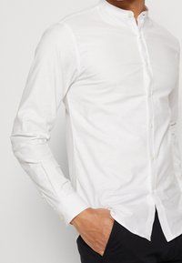 Witte blouse met lange mouwen en een mandarin-kraag, voorzien van een knoopsluiting aan de voorkant en een gladde stoftextuur, gedragen met de handen in de zakken.