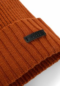 Gehaakte oranje beanie met een ribbelstructuur en een zwart leren logo-embleem. De stof lijkt zacht en stevig voor warmte.