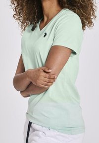T-shirt verde chiaro a scollo a V realizzata in tessuto morbido con maniche corte. Presenta un piccolo logo nero sul lato sinistro del petto e una vestibilità comoda.
