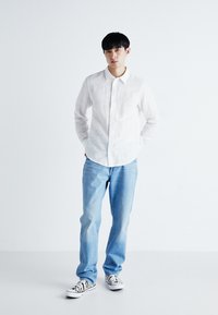 Camicia bianca a bottoni, jeans azzurri chiari e sneakers a righe bianche e nere. La camicia presenta un colletto e una leggera texture.