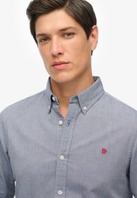 Młody mężczyzna z krótkimi ciemnymi włosami, ubrany w jasnoniebieską koszulę zapinaną na guziki z małym czerwonym haftowanym logo na piersi.