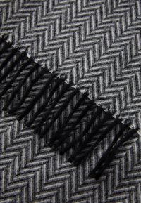 Écharpe en laine présentant un motif en chevrons noir et gris avec de longues franges noires le long d'un bord. Fini texturé et doux.