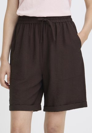 Kobieta nosząca ciemnobrązowe szorty z sznurkiem, załatanymi kieszeniami i podwiniętymi brzegami, w połączeniu z jasnoróżową bluzką.